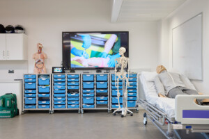 Clinical Skills Suite, showing skeletons, mannequin in a ward bed, a block of blue drawers, and a big television screen showing a procedure,