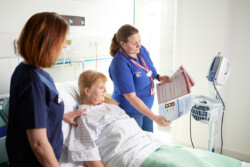 Student nurse showing papers to a patient in a ward bed, during a simulation. The academic is watching over.