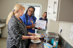 Student nurse showing a patient a document about how to make a sandwich, during a simulation. The patient is holding two slice of bread, with beans and butter on the kitchen side. The academic is in the background, observing.