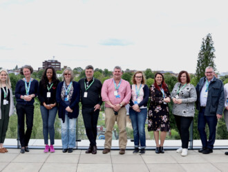17 delegates at the UON Sustainability Summit 2023 standing in a line, leaning against a balcony at the University of Northampton Waterside campus. The sky is cloudy, but it is bright.