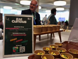 Two trays of food cups, one higher than the other. The lower one is the scramble power pot with Eat Curious Scrambled Negg, a vegan product. The sign next to it is from Eat Curious and says there is the Breakfast Wrap made by red onion marmalade, roast mushroom, vegan cheese and hash brown and the description of the Scambled Power Pot. In the background visitors can be seen getting food from other areas.