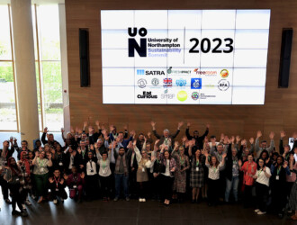 Group photo of all of the delegates, speakers, visitors, at the UON Sustainability Summit 2023. There are approximately 60 people in total and they are waving both or one hand up in excited motions. On the large screen taking up the entire back wall is the UON Sustainability Summit 2023 logo, along with a list of the sponsors.