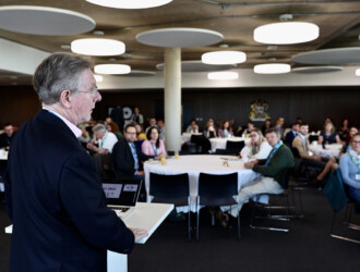 Speaker at the UON Sustainability Summit 2023, standing at the front table looking out towards tables with delegates watching them. There are approximately 6 tables seen full with guests.
