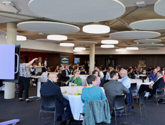 Large group of delegates at the UON Sustainability Summit 2023 sitting at approximately 8 tables in The Hide at the Learning Hub, in the background the breakfast buffet is seen.