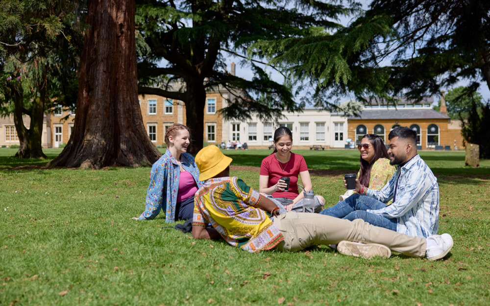 Five students sitting on the grass at Delapré Abbey, some are holding disposable coffee cups and another has a water bottle. There are trees behind them and in the distance the Abbey building. It is a sunny day, as some students are wearing sunglasses and hats.