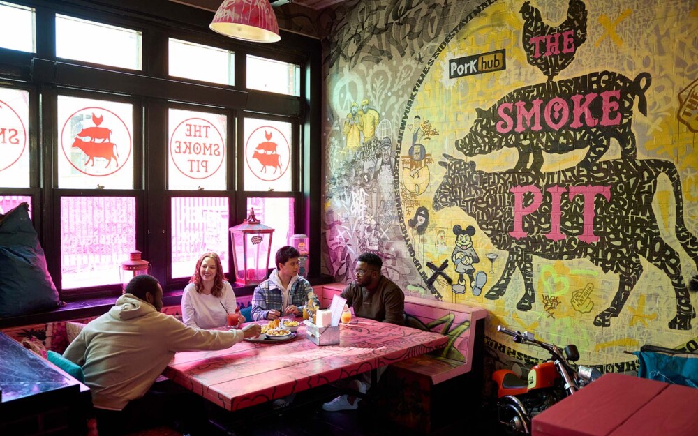 Internal view of the Smoke Pit restaurant where four students are sitting at a pink table, with pink benches in s square. On the wall is art of a chicken, pig, and cow, with graffiti style designs.