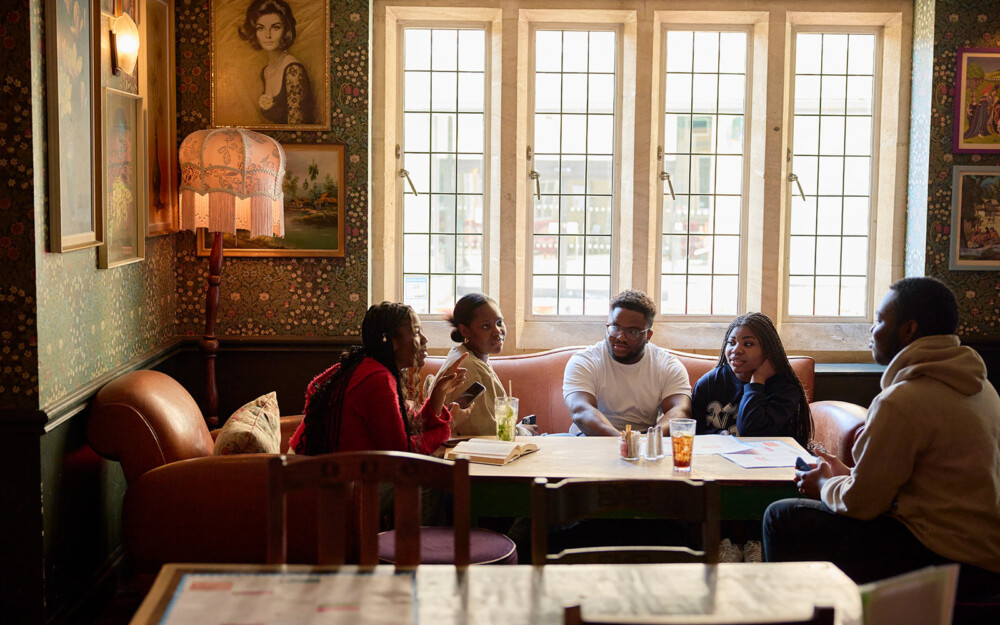 Five students sitting on leather sofa and armchair in the Zapato Lounge in Northampton. The windows are tall and the décor in the room includes paintings of portraits and landscapes, vibrant wallpaper of flowers over dark green.