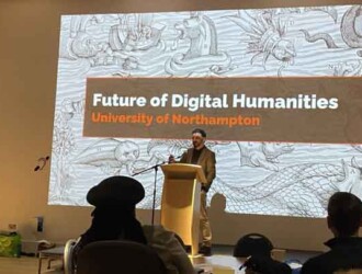 Chris Houghton from Gale International stands in front of a digital screen with a slide from his presentation called 'Future of Digital Humanities'