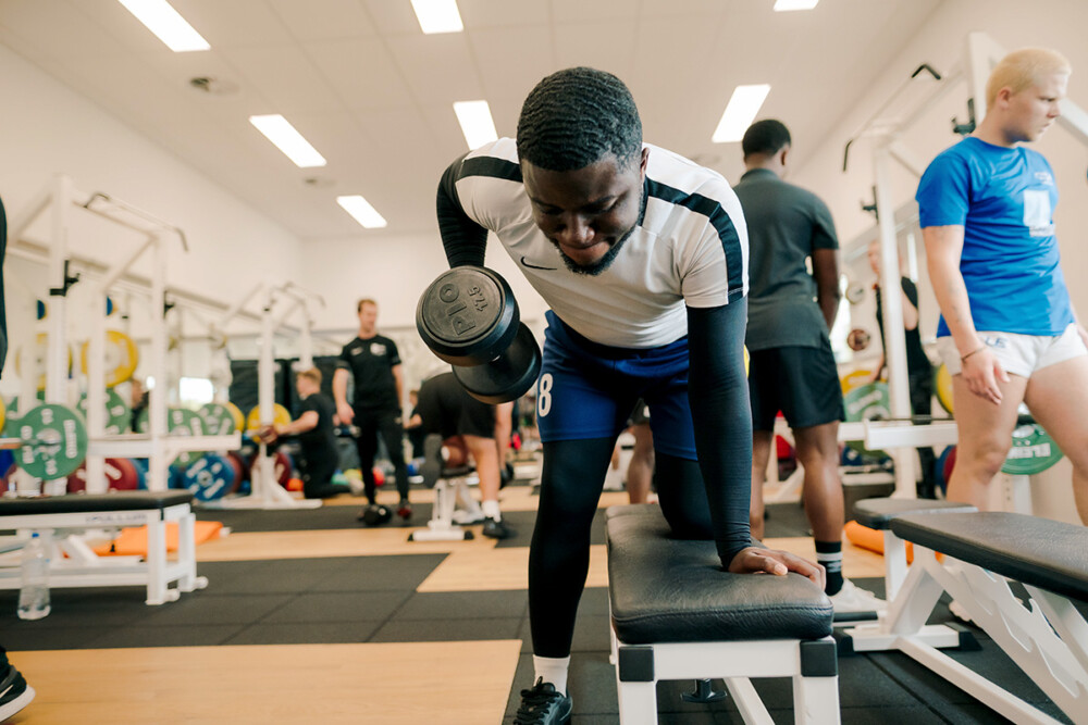 A student pulling a dumbbell towards them, while using their other hand to balance on the bench, doing a bent over dumbbell row. There are people behind also using the gym and gym facilities.