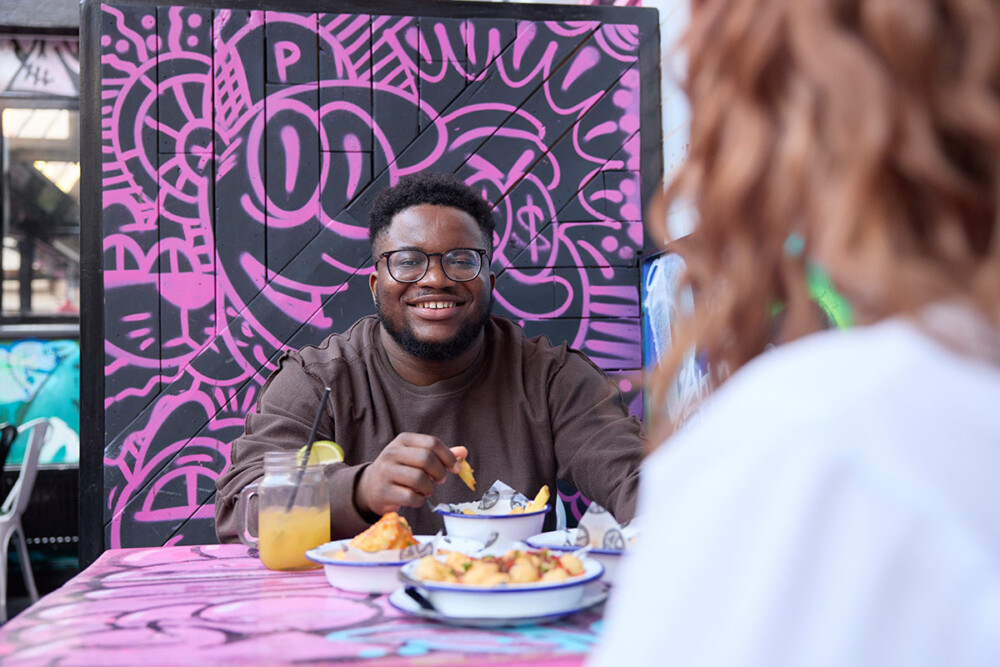 Two students at a restaurant, one is facing away from the camera and the other is looking towards it smiling, while holding a chip from their bowl of food. The decoration in the restaurant is pink graffiti.