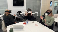A group of schoolchildren use virtual reality headsets in a classroom. They are sitting at a large white school table.