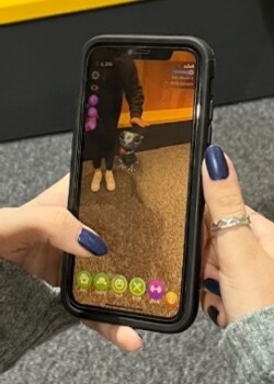A person is holding a smartphone in two hands - with the phone screen clearly visible. Extended reality figures appear on a smartphone screen