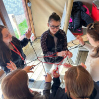 Five education students stand in a circle inside the forest school teepee, collaboratively working on a string collaboration project, with red and black strings forming intricate patterns between their hands.