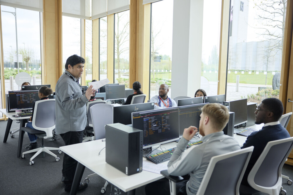 An academic holding papers while explaining a task to a group of students who are all sitting at specialist terminals in the Bloomberg Finance Lab. Large windows are in the background.