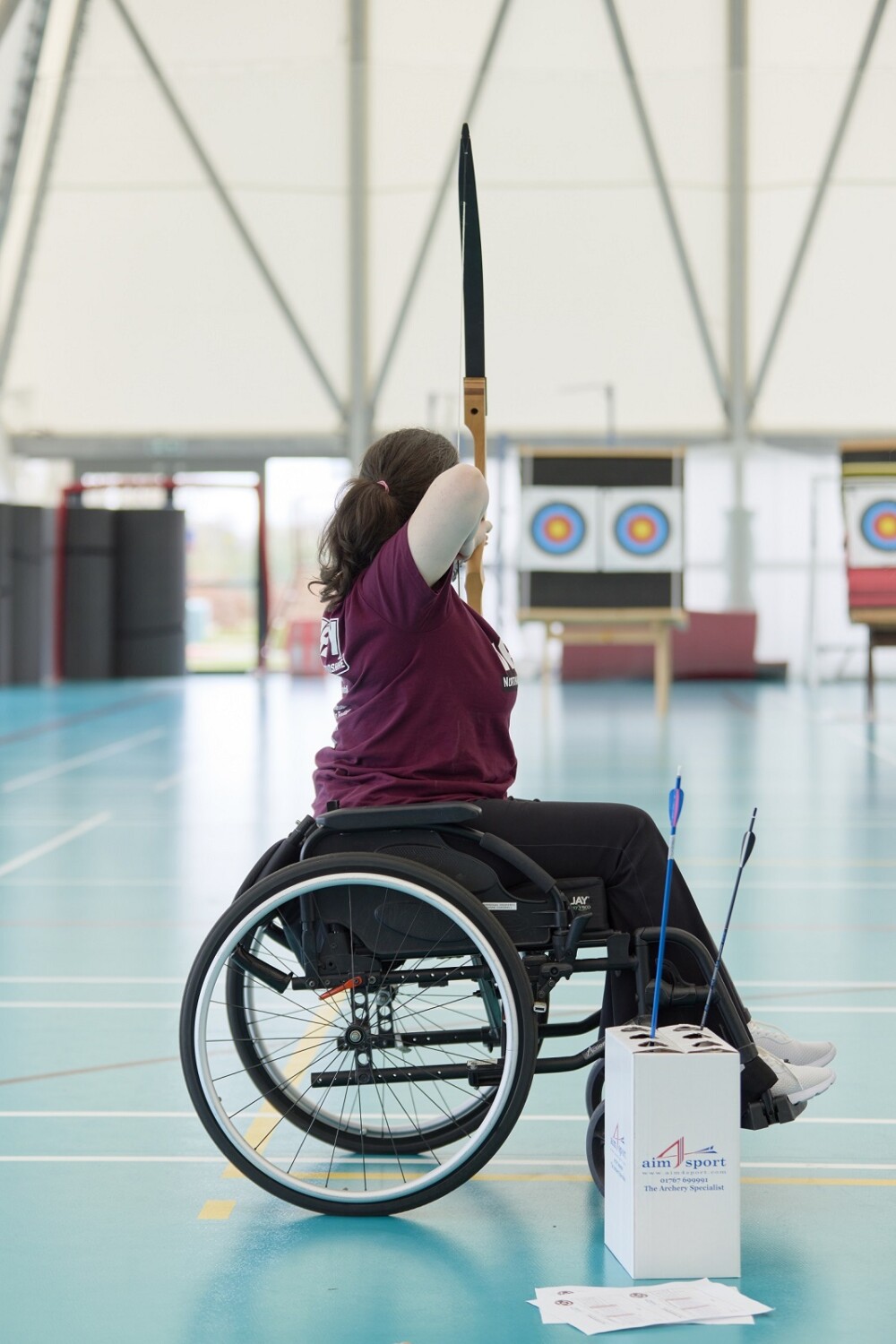 A student in a wheelchair aims a bow and arrow at an archery target at the other end of an indoor sports hall. She is facing away from the camera and wearing a maroon jersey. Spare arrows are resting in a cardboard holder by her feet.