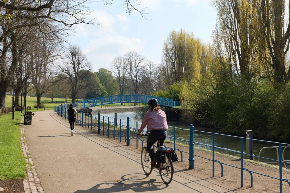 A person rides a bicycle and another jogs on a path alongside a river with trees and a blue footbridge in the background.