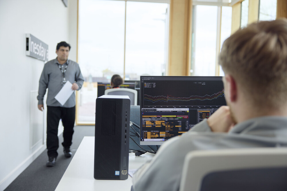 In the foreground, a student is using a specialist terminal during a class in the Bloomberg Finance Lab, while in the background, an academic walks around the classroom.