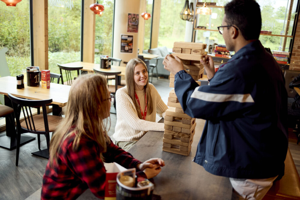 Three students play Jenga at a table in a restaurant. One stands to adjust a block, while the others watch. Behind them, tables and chairs are visible.