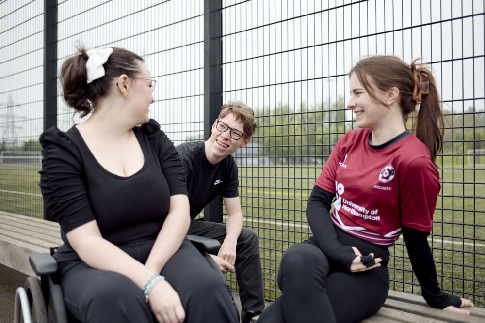 Three students are talking and smiling next to a black wire fence by a sports pitch. Two are seated on benches, and the third is seated in a wheelchair. Two are dressed in black, and the other is wearing a UON Student Union Archery Society uniform. They are outdoors on a cloudy day.