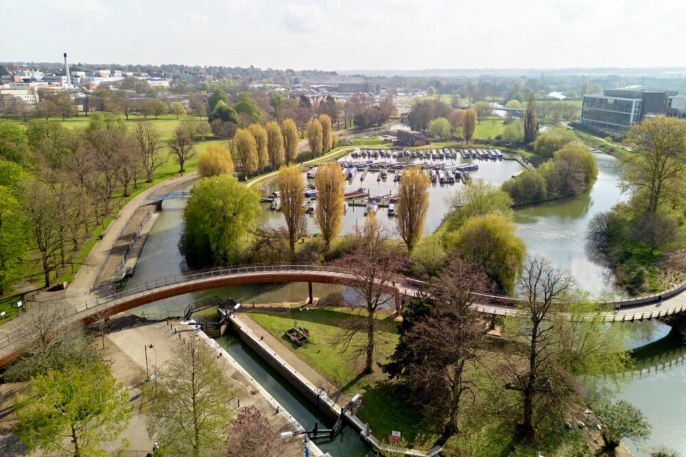 Aerial view of Northampton Marina and Becket’s Park. The landscape features a winding river, a wooden bridge, and a marina with boats. In the background, there are green trees and grassy areas, as well as Northampton town buildings.