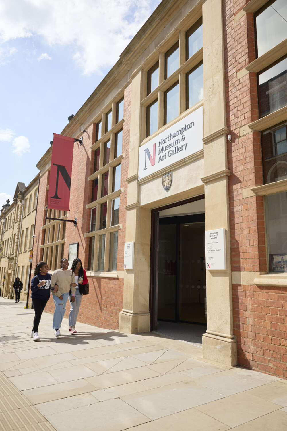 Three people walk past the entrance of Northampton Museum & Art Gallery, a red brick building with large windows.