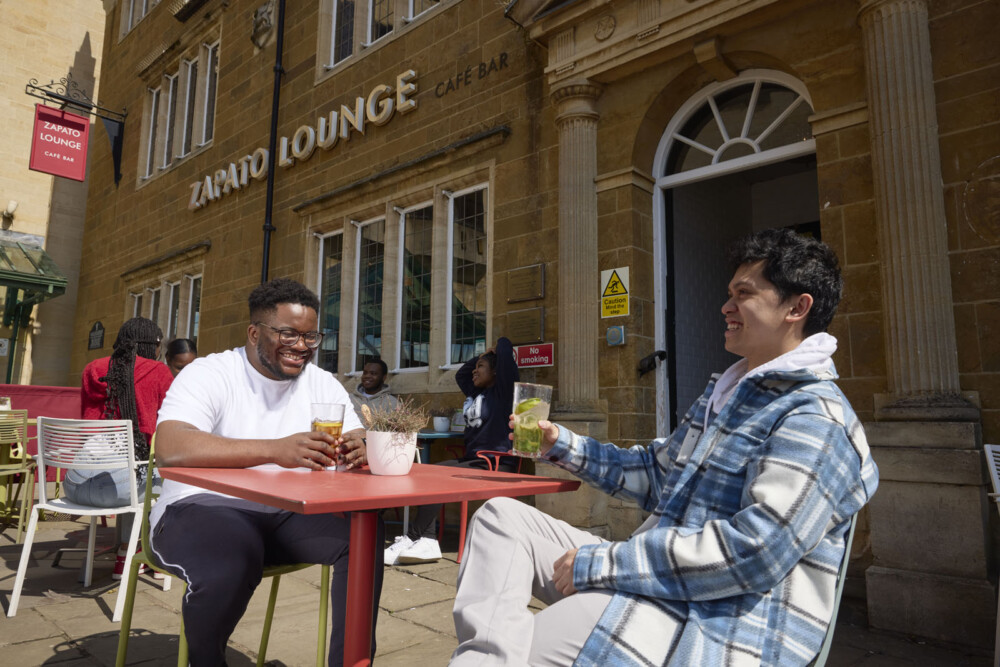 Two students sit at an outdoor café table, smiling and holding drinks. Other patrons are in the background, and the venue sign above the entrance reads ‘Zapato Lounge Café Bar’.