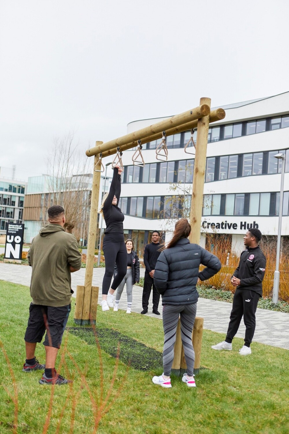 Image by Paul Michael Hughes Photography T 07790819111 UK E pmh@paulmichaelhughes.com W www.paulmichaelhughes.com A person wearing black top and leggings uses overhead rings on a piece of wooden outdoor gym equipment. She is being observed by five other students, all of whom are wearing either casual or athletic clothing. In the background, a large white building with ‘Creative Hub’ in large letters on the front is visible.