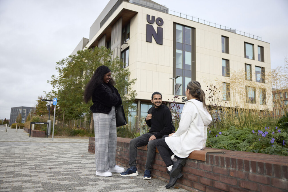 Three people talking outside a light-coloured modern building with a large UON logo. One is standing, while the others are sitting on a bench ledge. The standing woman is wearing check trousers and is holding a large black bag, and one of the seated students is wearing a light-coloured coat.