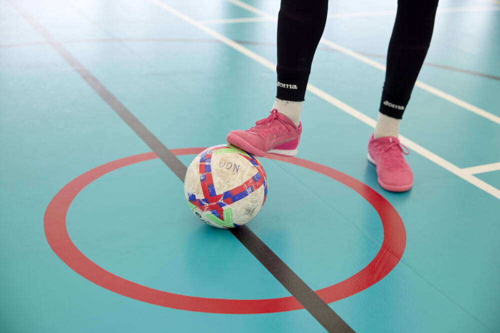A close-up of the legs and feet of a student wearing pink shoes and black leggings. They are standing with one foot on a football on an indoor court with blue flooring and red lines.