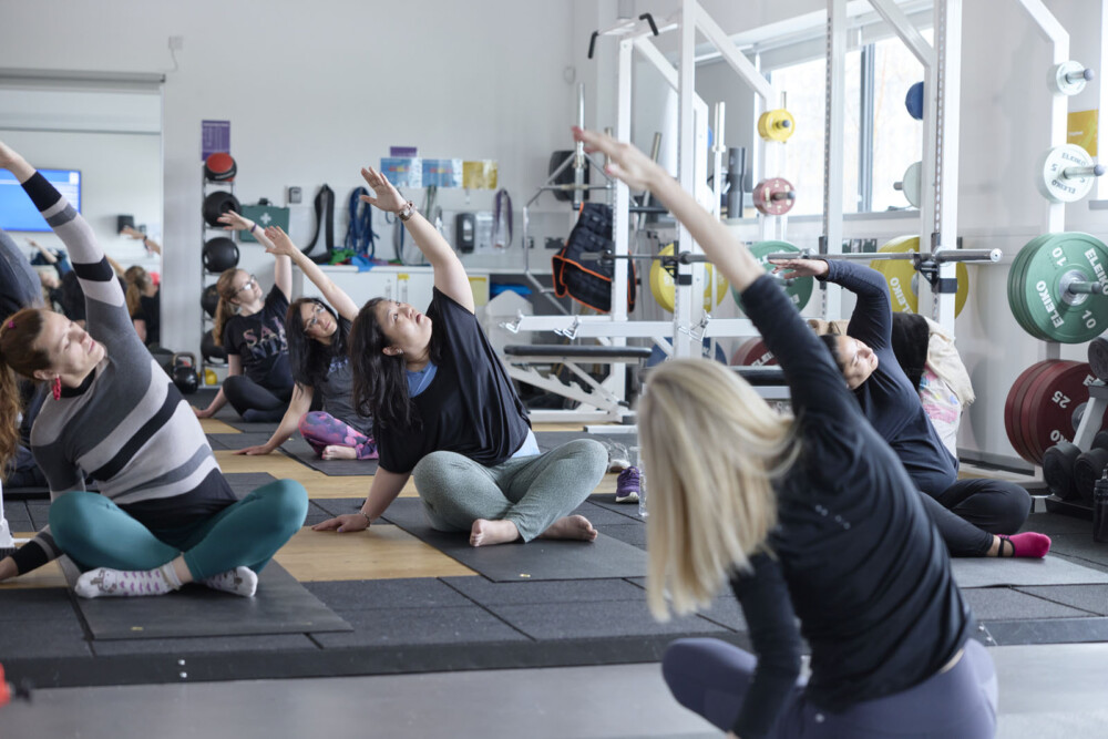 A group of people take part in a yoga class in a gym. They are seated and are performing side stretches. Behind them, various equipment including weights, rigs and benches are visible.