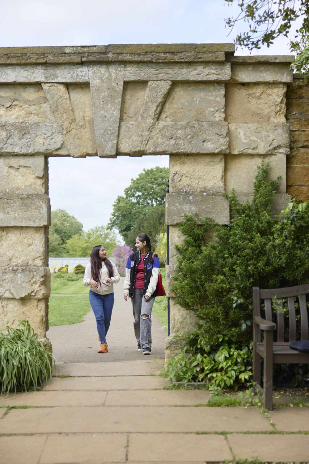 Two people walk side by side through a stone archway in a walled garden setting, with greenery and a bench nearby.