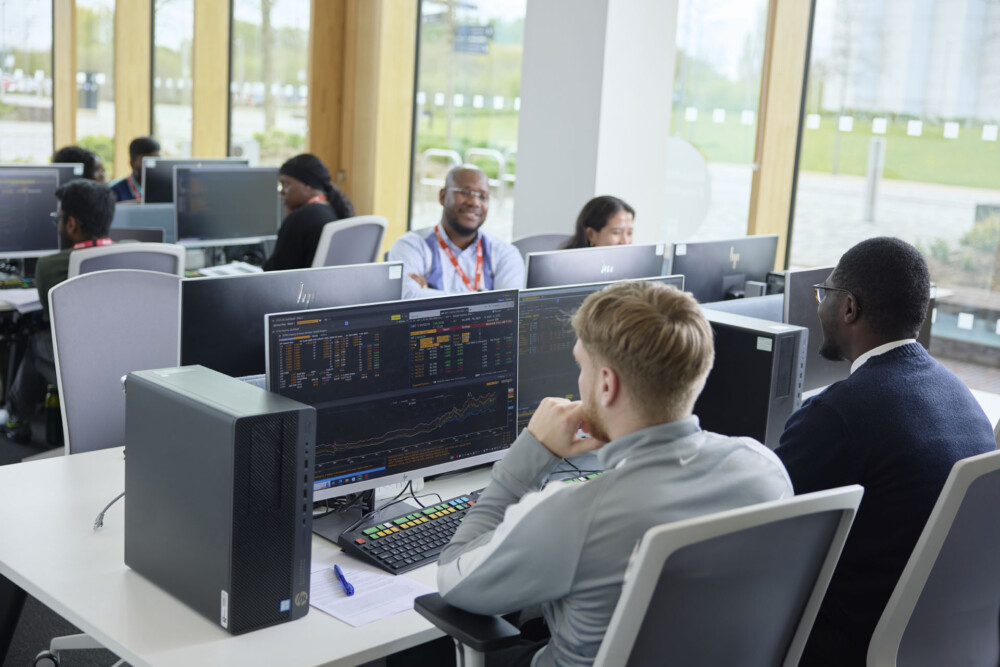 A group of students work on computers in a modern office with large windows. Screens display data and graphs.