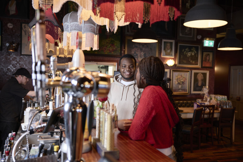 Two students stand at a bar counter, engaged in conversation. The setting is a cozy venue with eclectic art on the walls and a bartender working in the background.