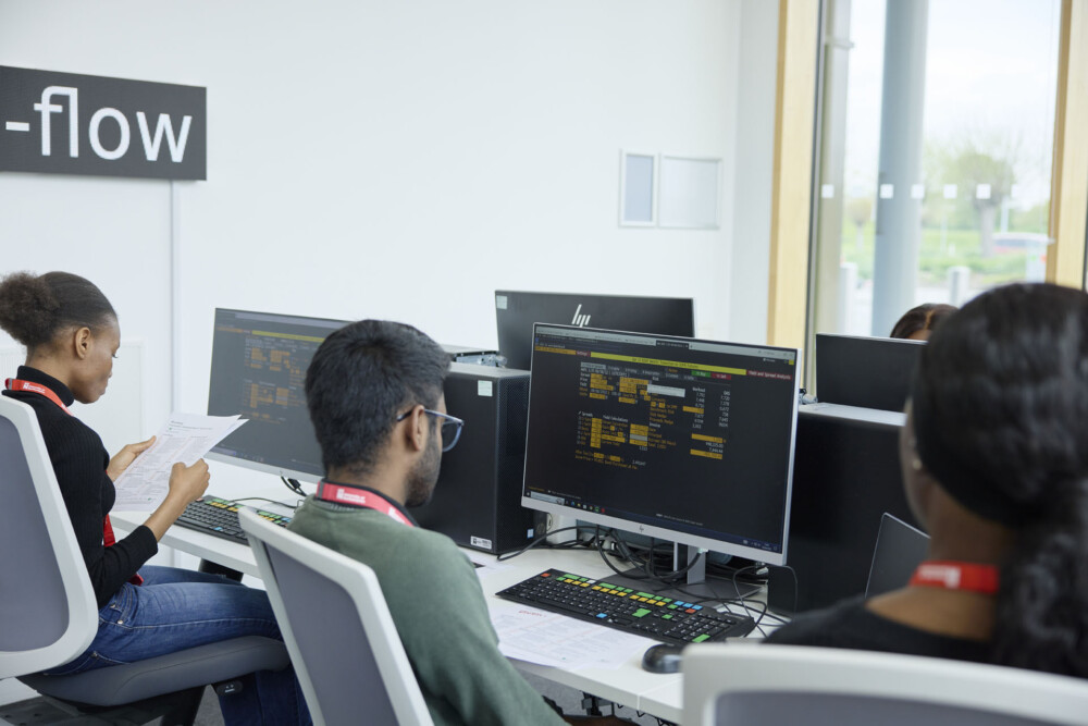 A group of students working on desktop computer terminals in the Bloomberg Finance Lab. On the wall, there is a large electronic sign.