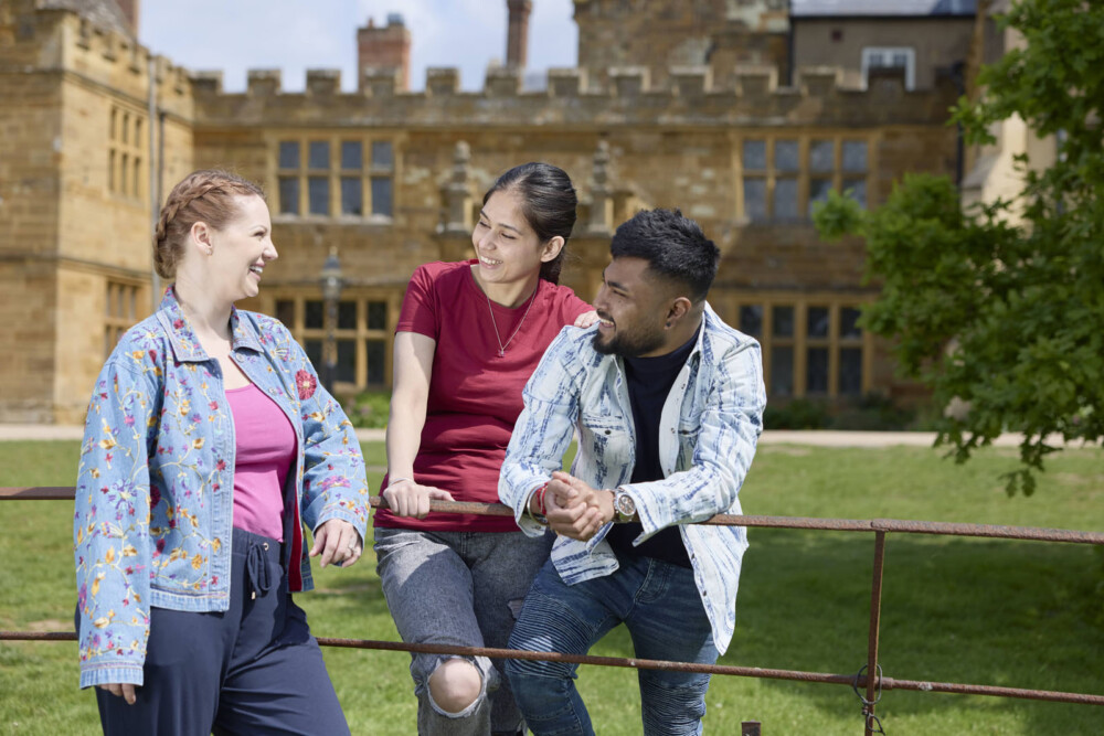 Three students chat and smile by a fence in front of a historical stone building on a sunny day.