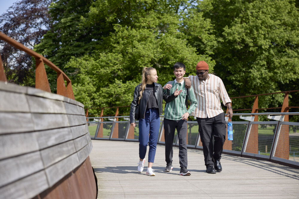 Three students walk and chat on a wooden bridge, surrounded by lush green trees. One of the students is wearing a beanie hat and carrying a large bottle of water, another wears a green and white striped top, and the other wears a black leather jacket.
