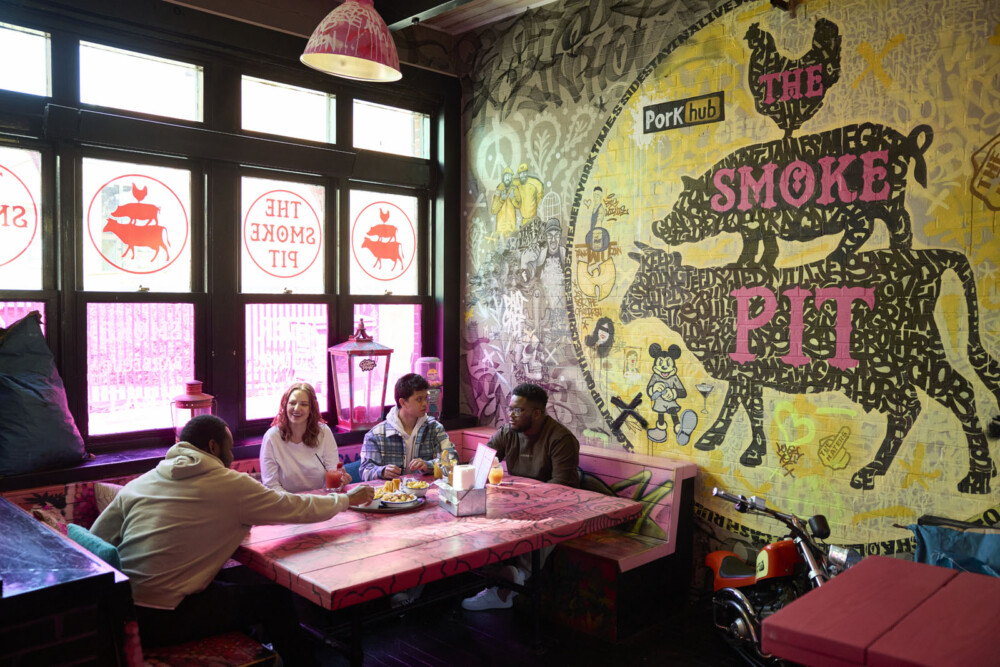 Four students sit around a table, sharing a meal. To the side, there is a large colourful mural of a cow, with a pig and chicken with ‘The Smoke Pit’ in large letters.