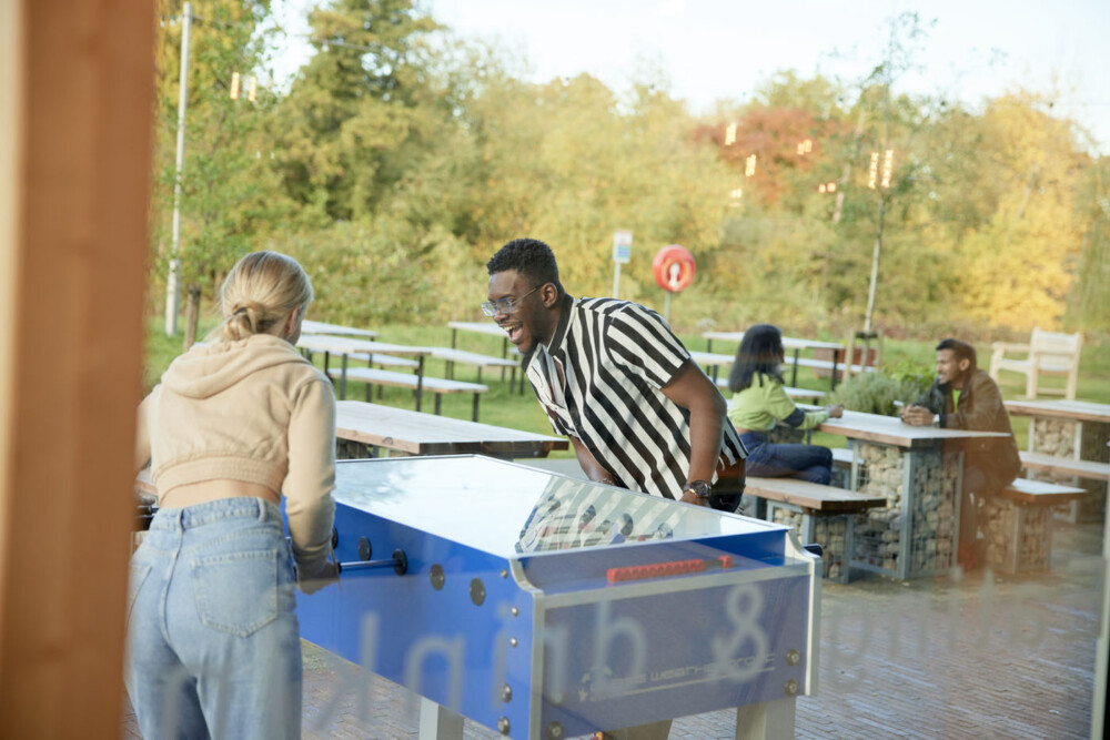 Two people playing table football outdoors. A woman in a cropped beige jacket and jeans faces a man in a striped shirt. In the background, a couple sit on one of several benches that are placed outside.