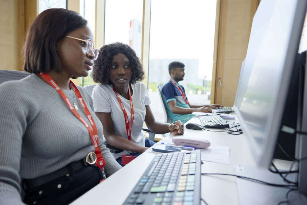 Two students using terminals and worksheets in the Bloomberg Finance Lab. In the background, another student works on a terminal. All are wearing red lanyards.
