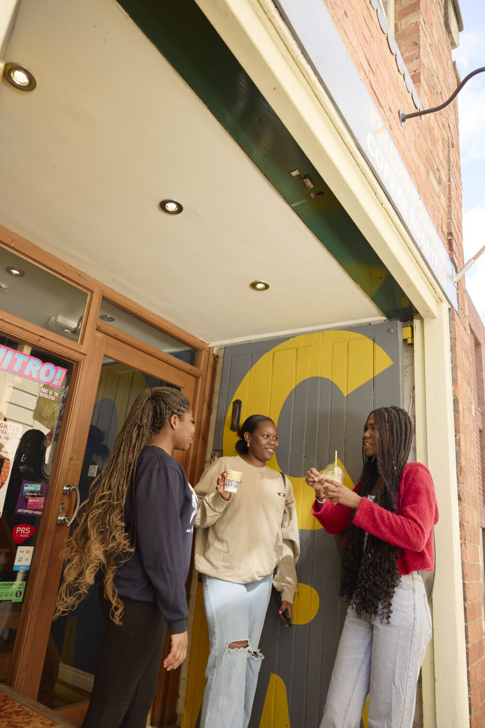 Three people stand outside a shop entrance, holding drinks and chatting. The entrance has vibrant yellow patterns.
