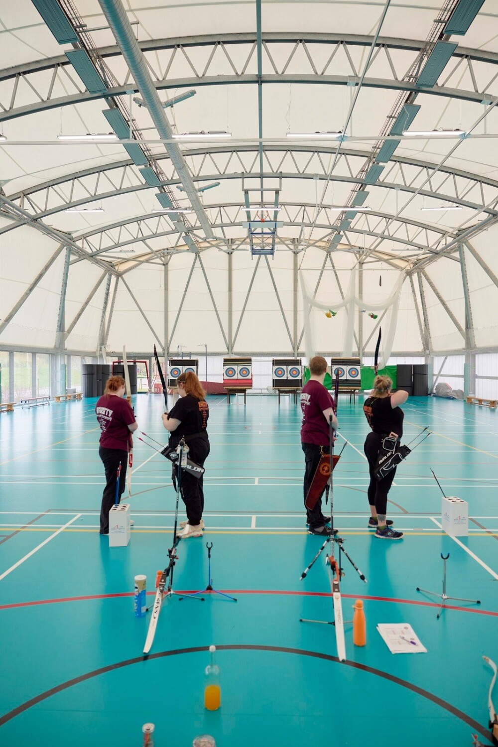 Four students take part in an archery competition. They have their backs to the camera and taking aim at targets at the other end of a large, bright sports hall. They are either wearing maroon or black tops, and three are carrying quivers.