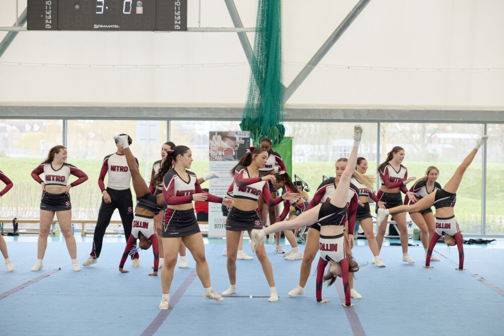 A female cheerleading team practicing a routine indoors, with members in coordinated maroon, black, and white uniforms, executing various poses and stunts on a blue mat.