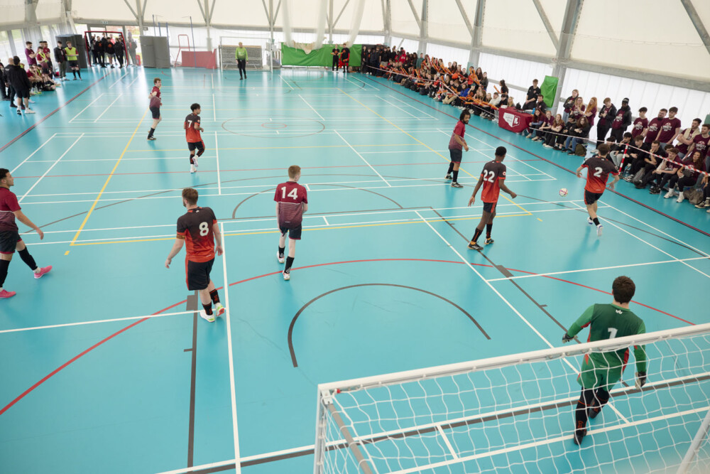 An indoor football match is in progress, with players in maroon and white or orange and black jerseys, which is taking place on a blue indoor pitch. The photo has been taken from a high angle above one of the goals. Spectators are visible on both sidelines.