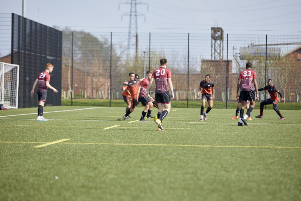 Two football teams – dressed in either maroon and white or orange and black jerseys – are playing in a green artificial turf field. The pitch is surrounded by a black wire fence and in the background, trees and buildings are visible.