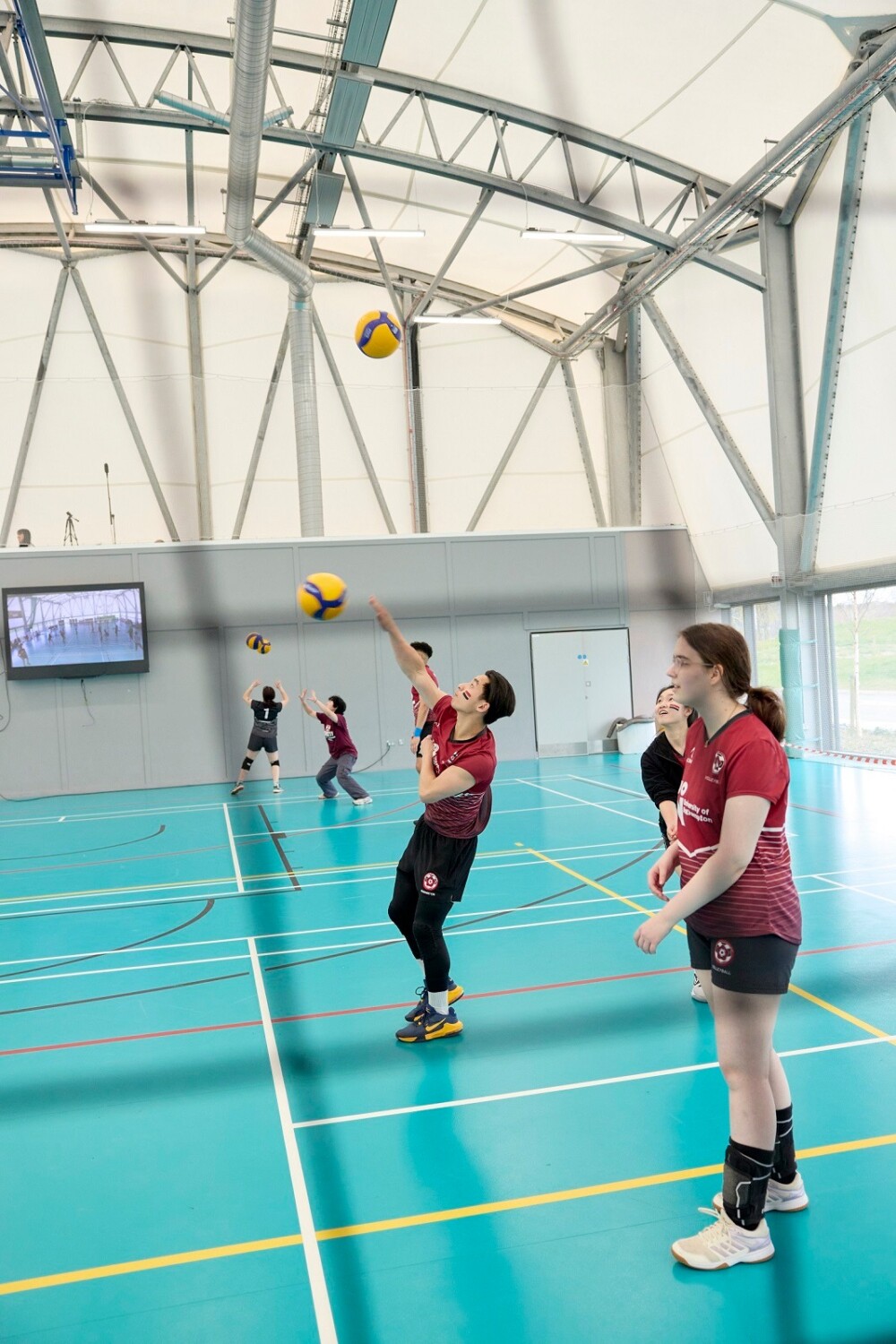 Students wearing maroon jerseys play volleyball on a blue court inside. One player is serving, while others prepare. There is a large wall-mounted screen in the background.