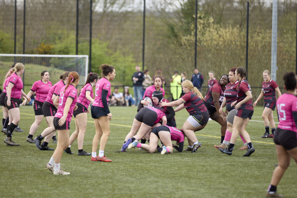 A women's rugby match in progress on an outside pitch. Players wearing either maroon with black stripes or pink jerseys are competing, with a group tackling for the ball while others observe. Spectators are visible in the background.