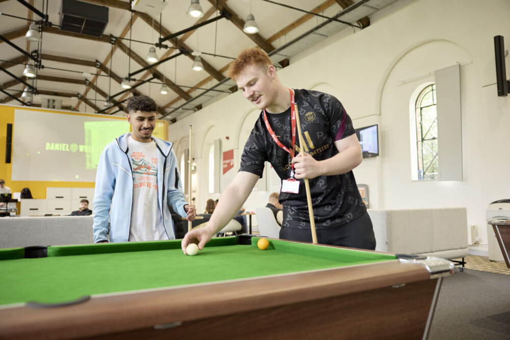 Two students play snooker in the Engine Shed. One student watches while the other lines up a shot with the cue ball. Both are casually dressed. Behind them, there are sofas and tables. The building has a high ceiling and is brightly-lit.