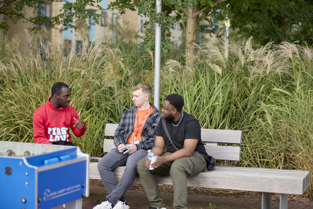 Three students sit chatting on a bench outside. Behind them, trees and greenery are visible in front of buildings. They are casually dressed, and one has a red lanyard. In the foreground is a table football table.