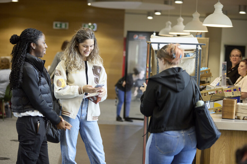Three students stand close to the counter of the Ground coffee shop. One is holding a packed sandwich and is looking down at her phone. In the background, two staff members stand behind the counter, and a person is looking inside a large fridge.
