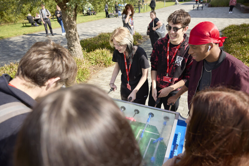 Group of students playing table football outside. They are casually dressed and wearing red lanyards. In the background, other people are walking or sitting on picnic benches or grass nearby. It is a sunny day.